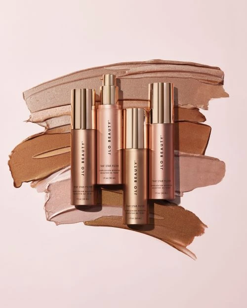 Four JLo Beauty serum bottles arranged on a soft pink surface with beige and brown cosmetic swatches for a beauty product shot.