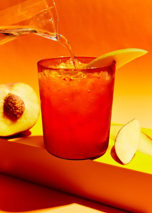 Pouring clear liquid into a red drink with ice cubes, garnished with mango, next to a peach on a yellow surface against orange background.
