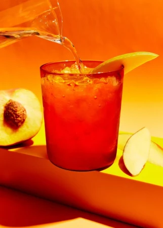Pouring clear liquid into a red drink with ice cubes, garnished with mango, next to a peach on a yellow surface against orange background.