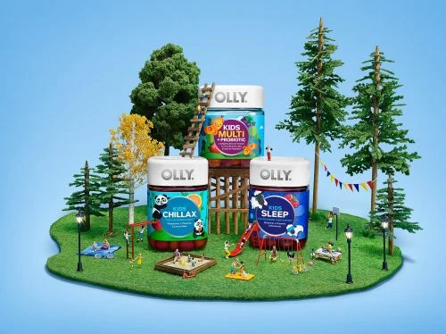 Three Olly Kids gummy vitamin bottles on a miniature playground with trees, lights, and playground equipment against a light blue backdrop.