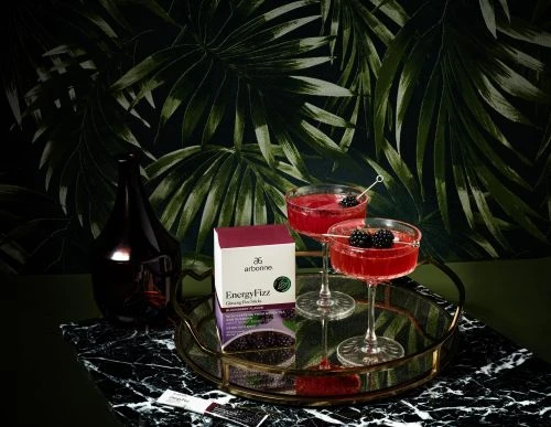 Two cocktails with blackberries on a gold tray with an Arbonne energy fizz box against a dark green floral wallpaper background.