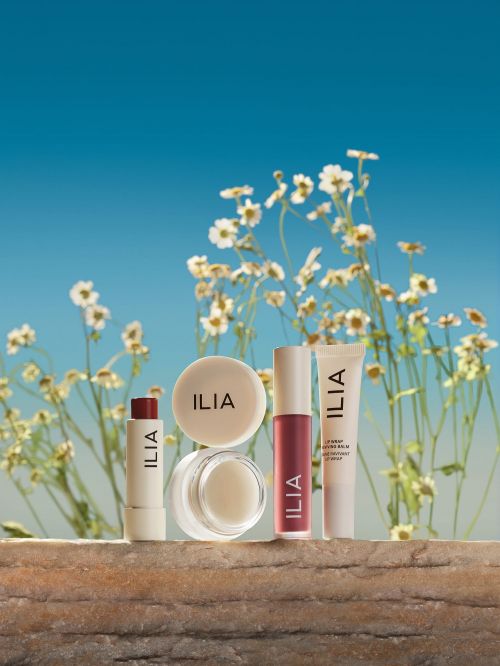 A collection of ILIA beauty products including lip balm, lip tint, and face powder arranged on a textured surface with a floral background.