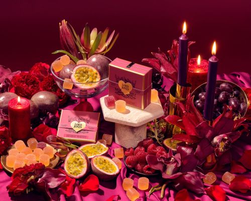 Still life with Lord Jones gummies, passion fruit, raspberries, grapes, candles, and flowers in shades of red and purple on a draped table.