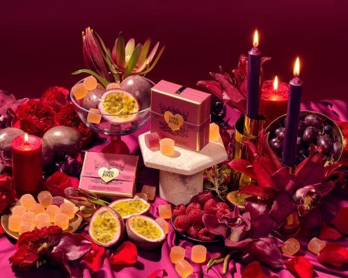 Still life with Lord Jones gummies, passion fruit, raspberries, grapes, candles, and flowers in shades of red and purple on a draped table.