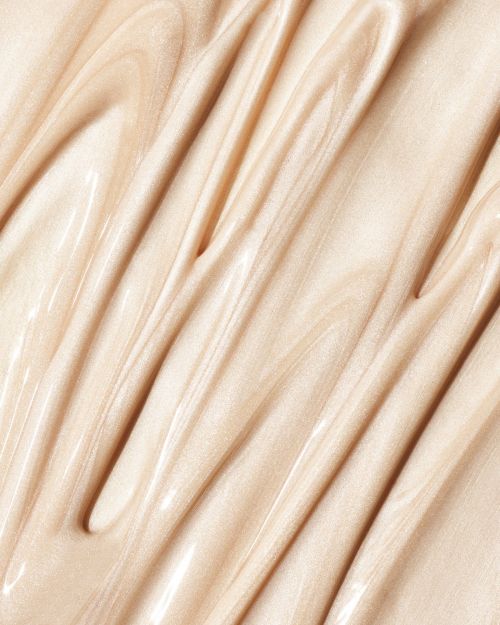 Close-up shot of a shiny, light beige cream, swirled and rippled to display its smooth, thick texture, and light-reflecting qualities.
