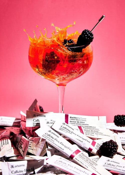Tall glass with a red and orange cocktail, blackberries, and splashes against a bright pink background, with scattered product packets.