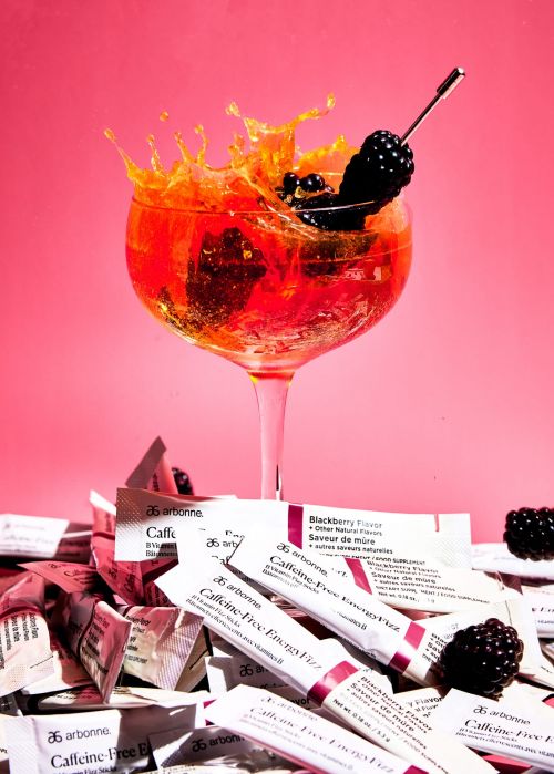 A cocktail glass with orange liquid, blackberries, and a splash, next to Arbonne Caffeine Free Energy Fizz packets, set against a pink background.