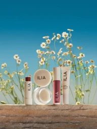 Ilia beauty products arranged on a stone ledge against wild white flowers and a clear blue sky