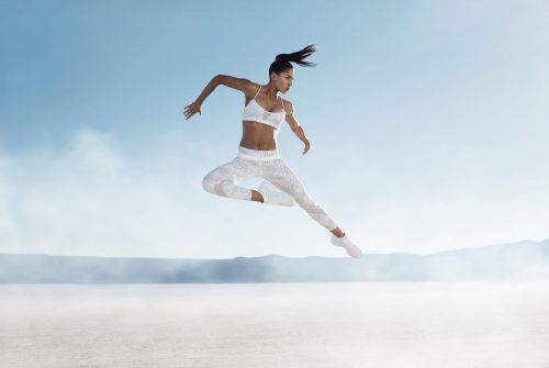 Athletic woman in white activewear mid-air, leaping against a clear blue sky, with a desert landscape in the background.