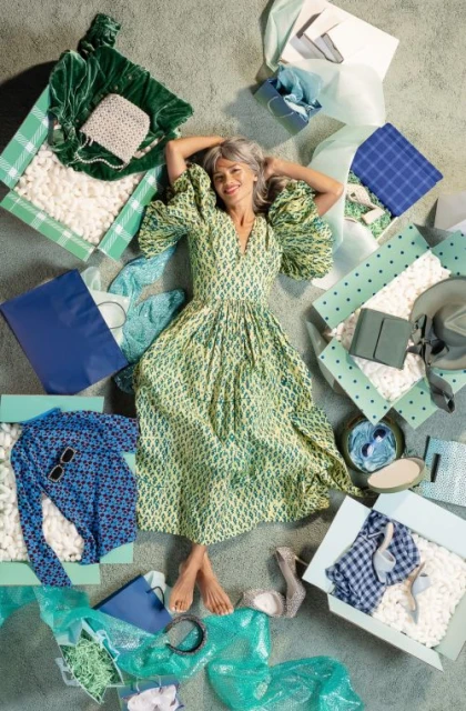 Woman in a green patterned dress lies on a carpet surrounded by opened boxes filled with clothes, shoes, handbags, and packing peanuts.