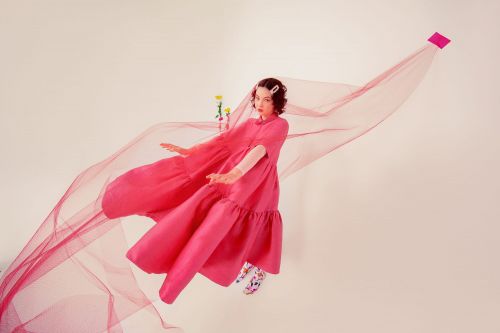 Young woman in pink dress with sheer fabric, floating against a white background. Fashion editorial with vibrant color and soft lighting.