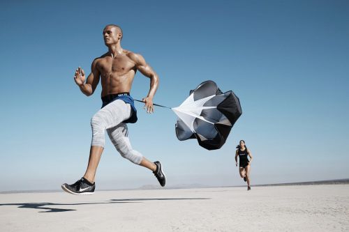 Muscular man runs on dry lakebed while pulling a parachute; another runner appears in the background, against a cloudless blue sky.
