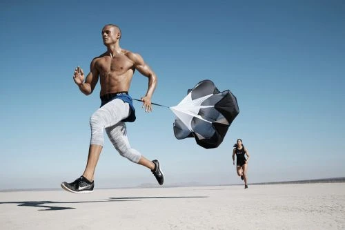 Muscular man runs on dry lakebed while pulling a parachute; another runner appears in the background, against a cloudless blue sky.