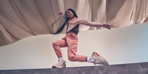 Athletic woman in pink outfit performs a dynamic pose with neutral-toned sneakers against a draped fabric backdrop in an active lifestyle shot.