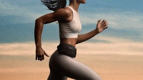 Athletic woman running outdoors wearing a sporty top and leggings with a fanny pack against a gradient sky background. Fitness.