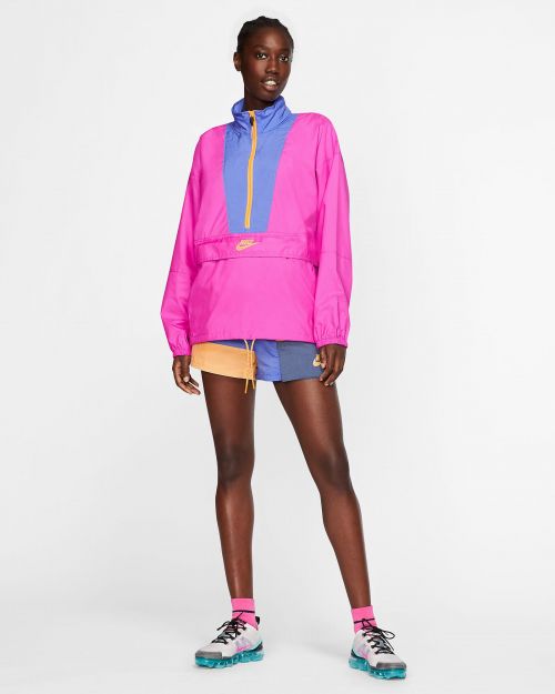 A young Black woman is standing in a photo studio wearing a pink and purple Nike jacket with matching accents on her shoes and socks.
