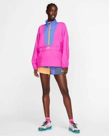 A young Black woman is standing in a photo studio wearing a pink and purple Nike jacket with matching accents on her shoes and socks.