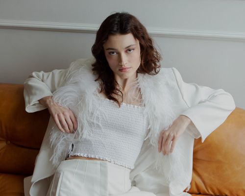 A poised young woman in a white top and pants, accented by a feathered blazer, is seated on a brown sofa against a neutral wall.