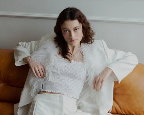 A poised young woman in a white top and pants, accented by a feathered blazer, is seated on a brown sofa against a neutral wall.