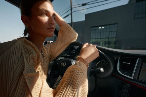 A woman sits in the driver's seat of a modern car, bathed in sunlight, with a city building visible in the background.