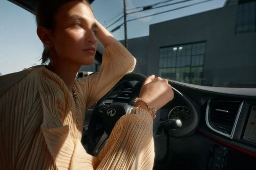 A woman sits in the driver's seat of a modern car, bathed in sunlight, with a city building visible in the background.