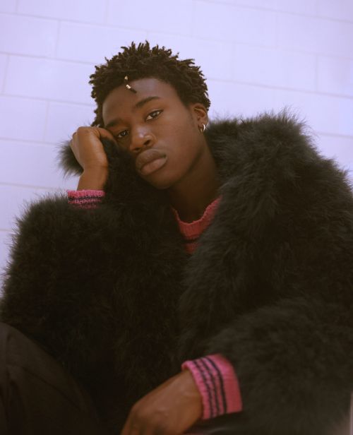 Young Black man in a feathered jacket poses indoors against a tiled wall, with soft lighting highlighting his features and outfit.