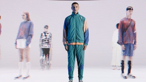 Man in a teal tracksuit stands in a studio with blurred figures, illuminated by bright, even lighting for a fashion or editorial shoot.