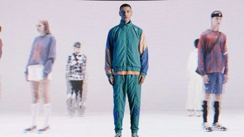 Man in a teal tracksuit stands in a studio with blurred figures, illuminated by bright, even lighting for a fashion or editorial shoot.