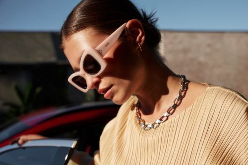 Brunette woman with sunglasses and necklace posing near a car, wearing a pleated top, outdoors in daytime.