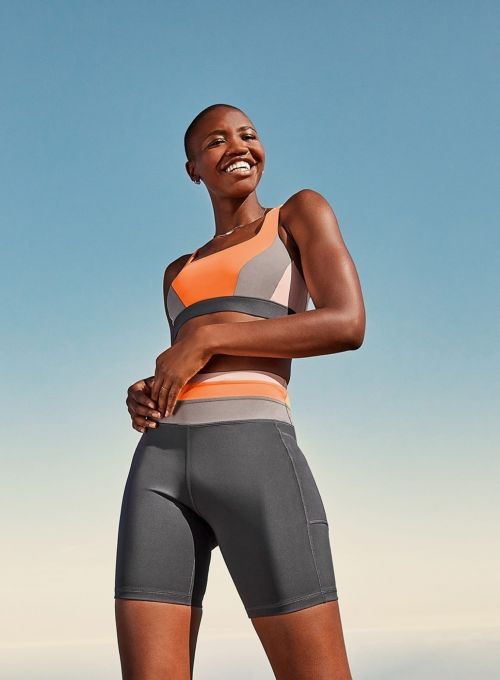 Smiling woman in sports bra and bike shorts stands against blue sky, looking up, posing for a fitness or activewear lifestyle shot.