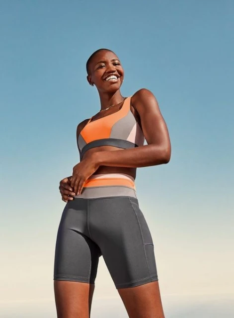 Smiling woman in sports bra and bike shorts stands against blue sky, looking up, posing for a fitness or activewear lifestyle shot.