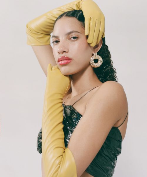 Portrait of a young woman with dark curly hair, red lipstick, and yellow leather gloves, posing in front of a neutral background.