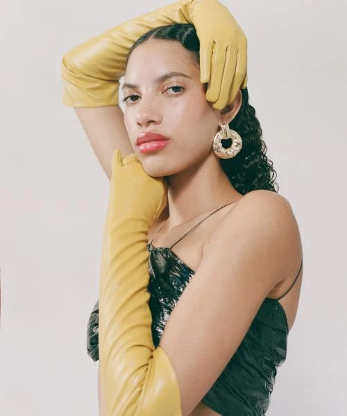 Portrait of a young woman with dark curly hair, red lipstick, and yellow leather gloves, posing in front of a neutral background.