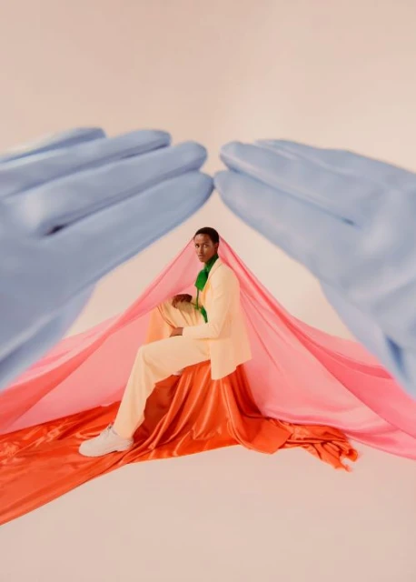 Man in peach suit poses with blue-gloved hands in studio under pink and orange fabric, wearing white sneakers and green scarf.