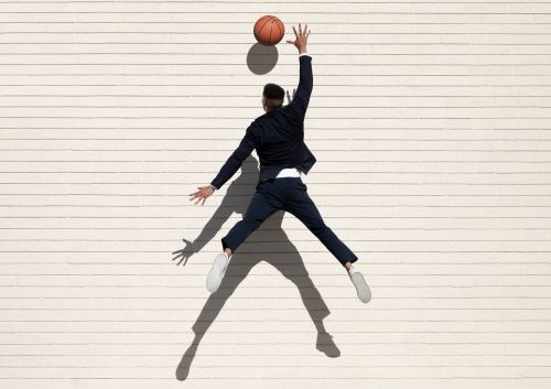 Young man in a suit jumps to catch a basketball against a white horizontal-plank wall, casting a long shadow on the surface.