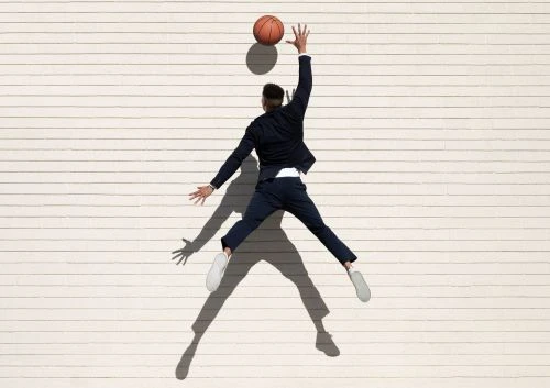 Young man in a suit jumps to catch a basketball against a white horizontal-plank wall, casting a long shadow on the surface.