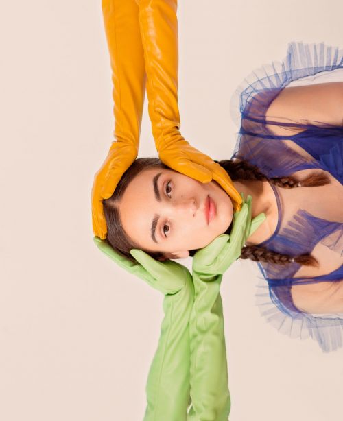 Portrait of a woman with light skin, brown hair, blue dress, and yellow and green opera gloves framing her face against a neutral background.