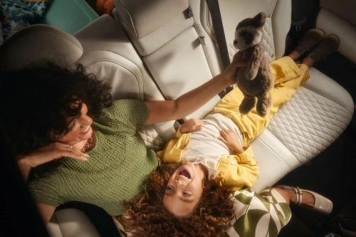 Mother and child lie in car seat, playing with a stuffed animal toy. The woman wears a green knit top; the child wears yellow overalls.