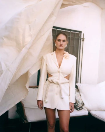 Portrait of a woman in a cream-colored blazer dress with cutouts and a rope belt, posing against a white wall with a window.