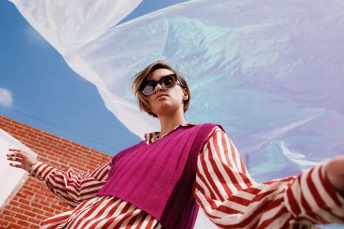 Woman with sunglasses poses outdoors wearing striped shirt and magenta vest against blue sky and blowing translucent plastic.