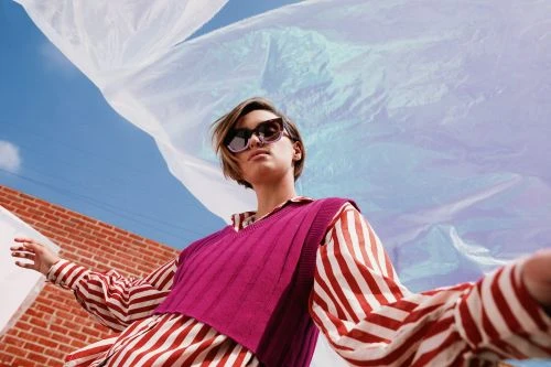 Woman with sunglasses poses outdoors wearing striped shirt and magenta vest against blue sky and blowing translucent plastic.
