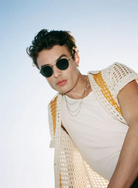 Man in sunglasses poses in a low-angle shot wearing a white t-shirt and crocheted cardigan against a bright blue sky backdrop.