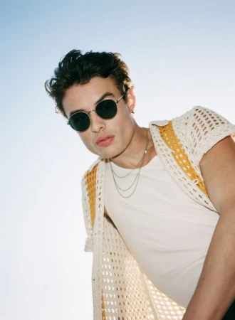Man in sunglasses poses in a low-angle shot wearing a white t-shirt and crocheted cardigan against a bright blue sky backdrop.