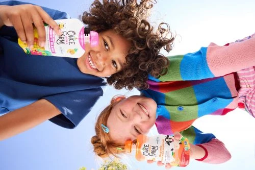 Two happy children smile up at the camera holding bottles of Once Upon a Farm organic juice, clear blue sky in the background.
