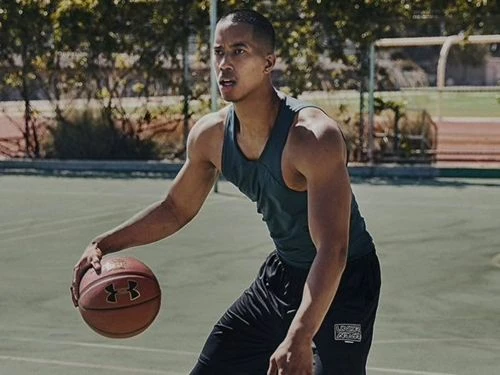 Athletic man in tank top dribbles basketball on outdoor court on sunny day, showing activewear brand logo on shorts and basketball.