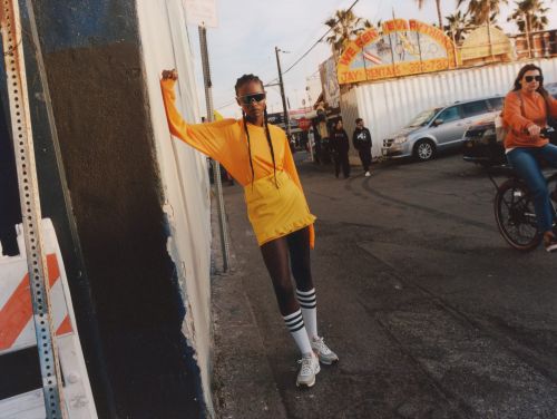 Stylish woman in yellow outfit poses on urban street with palm trees and passing traffic in soft daylight, fashion-forward and trendy.