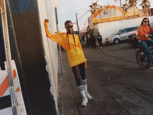 Stylish woman in yellow outfit poses on urban street with palm trees and passing traffic in soft daylight, fashion-forward and trendy.