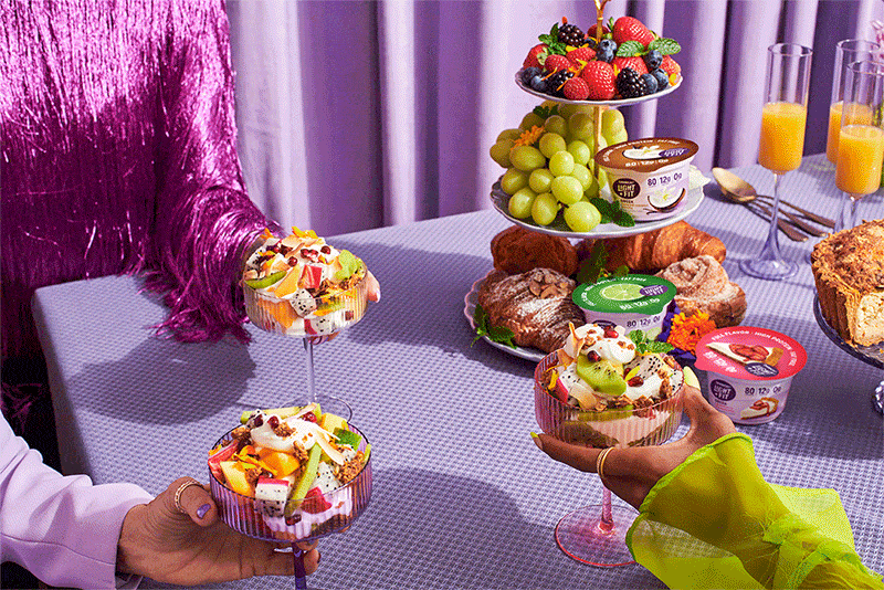 Eye-level shot of a tiered dessert stand and parfait glasses, surrounded by vibrant fruits, pastries, yogurt containers, and refreshing orange juice.