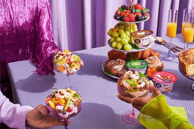 Eye-level shot of a tiered dessert stand and parfait glasses, surrounded by vibrant fruits, pastries, yogurt containers, and refreshing orange juice.