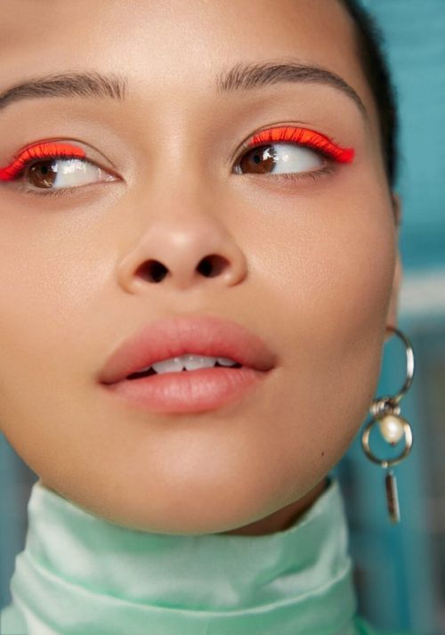 Close-up of a young woman with striking neon orange eyeliner. The makeup highlights her clear skin and light pink lips in a beauty shot.